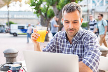 Mature Man Working On Laptop Drinking Orange Juice At Sidewalk Cafe