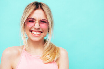 Closeup portrait of young beautiful smiling female in trendy summer hipster clothes. Sexy carefree woman posing on blue background in studio. Positive model winks. In sunglasses