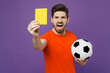 Young fan man he wears orange t-shirt angry at football sport team hold in hand soccer ball watch tv live stream hold yellow card propose player retire from field isolated on plain purple background.