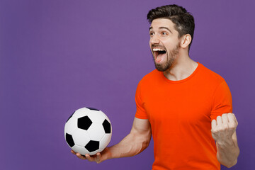 Wall Mural - Young fun exultant happy fan man he 20s wearing orange t-shirt cheer up support football sport team hold in hand soccer ball watch tv live stream do winner gesture isolated on plain purple background.