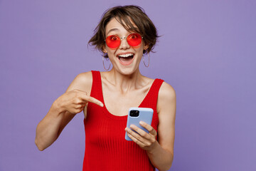 Wall Mural - Young shocked surprised happy fun cool woman 20s she wear red tank shirt eyeglasses hold in hand use point index finger on mobile cell phone isolated on plain purple color backround studio portrait