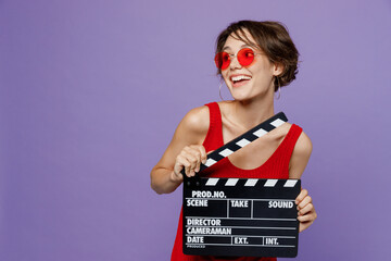 Wall Mural - Young smiling woman 20s she wear red tank shirt eyeglasses look aside on workspace holding classic black film making clapperboard isolated on plain purple backround studio. People lifestyle concept.