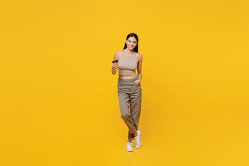 Wall Mural - Full body young latin woman 30s she wear basic beige tank shirt hold takeaway delivery craft paper brown cup coffee to go isolated on plain yellow backround studio portrait. People lifestyle concept