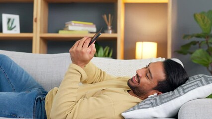 Aufkleber - Relaxed man texting on his phone and listening to music in his earbuds while lying on the sofa. Happy male browsing social media on an online app and smiling while relaxing on the couch at home