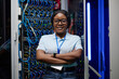© AnnaStills - Portrait of African female engineer smiling at camera standing with her arms crossed in data server with cables in rack in background