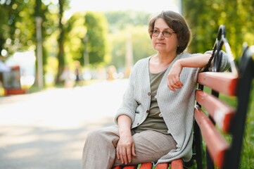 Canvas Print - Elegant elderly woman in the shirt is sitting on the bench in a park on a warm day