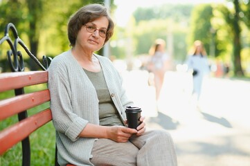 Poster - Elegant elderly woman in the shirt is sitting on the bench in a park on a warm day