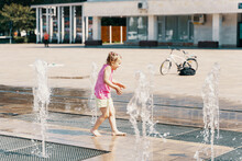 Little Girl Playing With Small Fountains In The Town Square On A Hot Summer Day