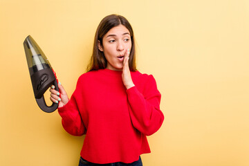 Wall Mural - Young caucasian woman holding a hand vacuum cleaner isolated on yellow background is saying a secret hot braking news and looking aside