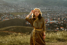 Young Mongolian Woman Wears Traditional Golden Deel Dress. Ulaanbaatar In Background At Sunset.
