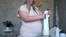 Happy Pregnant Woman With Big Belly Holding Baby Clothes In Hands At Home