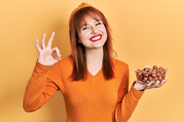 Canvas Print - Redhead young woman holding hazelnuts doing ok sign with fingers, smiling friendly gesturing excellent symbol