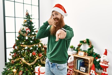 Canvas Print - Redhead man with long beard wearing christmas hat by christmas tree smiling doing talking on the telephone gesture and pointing to you. call me.