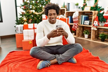 Sticker - Young african american man using smartphone sitting on the floor by christmas tree at home.