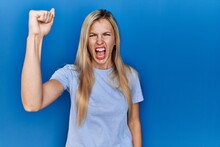 Beautiful Blonde Woman Wearing Casual T Shirt Over Blue Background Angry And Mad Raising Fist Frustrated And Furious While Shouting With Anger. Rage And Aggressive Concept.