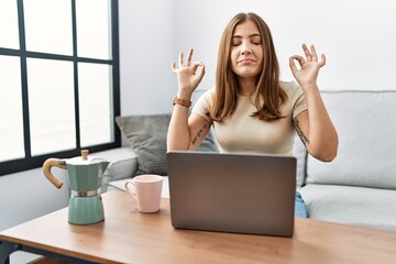 Canvas Print - Young brunette woman using laptop at home drinking a cup of coffee relax and smiling with eyes closed doing meditation gesture with fingers. yoga concept.