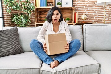 Canvas Print - Young hispanic woman sitting on the sofa checking delivery package angry and mad screaming frustrated and furious, shouting with anger. rage and aggressive concept.