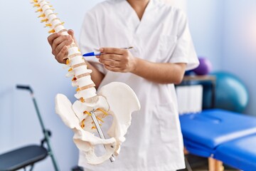 Poster - Young latin woman wearing physiotherapist uniform pointing to anatomical model of vertebral column at physiotherapy clinic