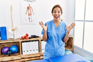 Canvas Print - Middle age hispanic physiotherapist woman working at pain recovery clinic celebrating crazy and amazed for success with arms raised and open eyes screaming excited. winner concept