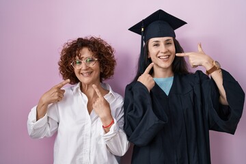 Wall Mural - Hispanic mother and daughter wearing graduation cap and ceremony robe smiling cheerful showing and pointing with fingers teeth and mouth. dental health concept.