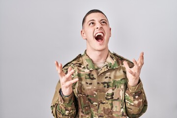 Canvas Print - Young man wearing camouflage army uniform crazy and mad shouting and yelling with aggressive expression and arms raised. frustration concept.