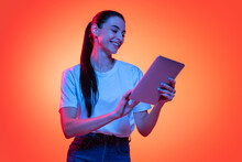 Portrait Of Young Emotional Girl, Student In White T-shirt Isolated On Orange Color Background In Neon Light. Concept Of Beauty, Fashion, Emotions