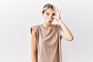 Wall Mural - Young blonde woman standing over isolated background doing ok gesture shocked with surprised face, eye looking through fingers. unbelieving expression.