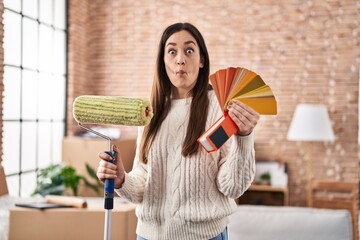 Poster - Young brunette woman holding roller painter and paint samples making fish face with mouth and squinting eyes, crazy and comical.