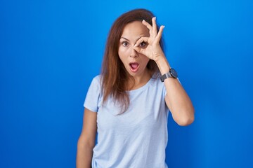 Poster - Brunette woman standing over blue background doing ok gesture shocked with surprised face, eye looking through fingers. unbelieving expression.