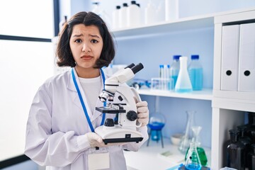 Canvas Print - Young hispanic woman working at scientist laboratory holding microscope skeptic and nervous, frowning upset because of problem. negative person.