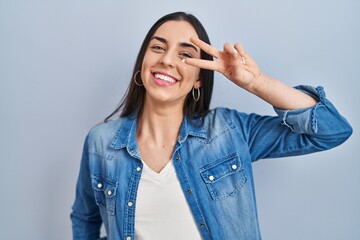 Wall Mural - Hispanic woman standing over blue background doing peace symbol with fingers over face, smiling cheerful showing victory