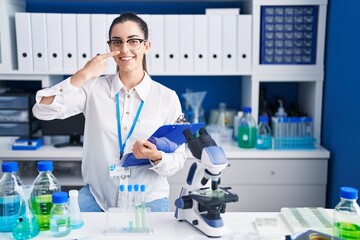 Poster - Young brunette woman working at scientist laboratory pointing with hand finger to face and nose, smiling cheerful. beauty concept