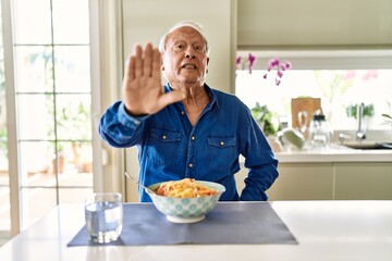 Sticker - Senior man with grey hair eating pasta spaghetti at home doing stop sing with palm of the hand. warning expression with negative and serious gesture on the face.