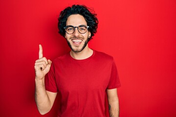 Canvas Print - Handsome hispanic man wearing casual t shirt and glasses showing and pointing up with finger number one while smiling confident and happy.