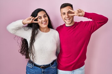 Sticker - Young hispanic couple standing over pink background doing peace symbol with fingers over face, smiling cheerful showing victory