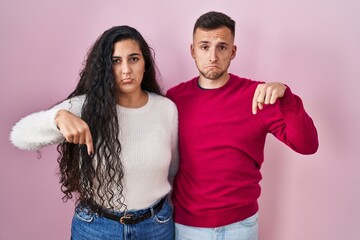 Sticker - Young hispanic couple standing over pink background pointing down looking sad and upset, indicating direction with fingers, unhappy and depressed.