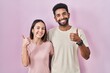 Young hispanic couple together over pink background doing happy thumbs up gesture with hand. approving expression looking at the camera showing success.