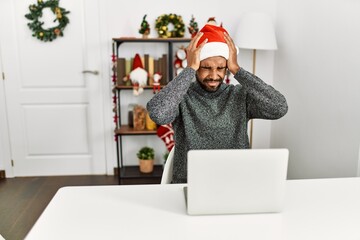 Poster - Young hispanic man with beard wearing christmas hat using laptop suffering from headache desperate and stressed because pain and migraine. hands on head.