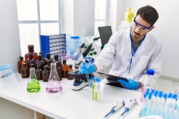 Wall Mural - Young hispanic man wearing scientist uniform writing on clipboard working at laboratory