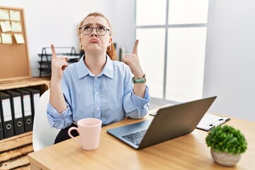 Wall Mural - Young redhead woman working at the office using computer laptop pointing up looking sad and upset, indicating direction with fingers, unhappy and depressed.