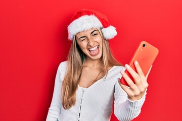 Poster - Beautiful hispanic woman wearing christmas hat using smartphone winking looking at the camera with sexy expression, cheerful and happy face.