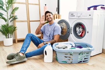 Sticker - Young hispanic man putting dirty laundry into washing machine waiving saying hello happy and smiling, friendly welcome gesture