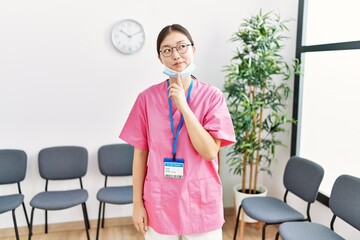 Wall Mural - Young asian nurse woman at medical waiting room thinking concentrated about doubt with finger on chin and looking up wondering