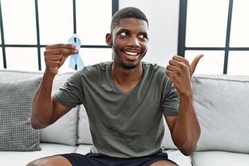 Poster - Young african american man holding blue ribbon sitting on the sofa at home pointing thumb up to the side smiling happy with open mouth