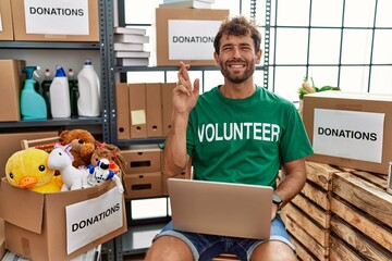 Poster - Young handsome man wearing volunteer t shirt using laptop gesturing finger crossed smiling with hope and eyes closed. luck and superstitious concept.