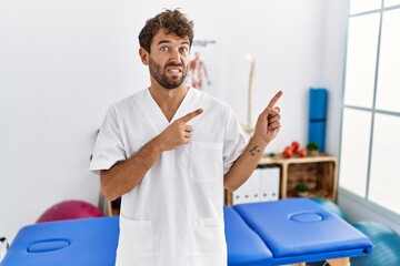 Canvas Print - Young handsome physiotherapist man working at pain recovery clinic pointing aside worried and nervous with both hands, concerned and surprised expression