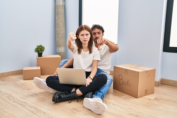 Sticker - Young couple using laptop at new home looking unhappy and angry showing rejection and negative with thumbs down gesture. bad expression.