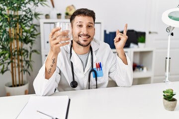 Canvas Print - Young doctor holding glass of water smiling happy pointing with hand and finger to the side