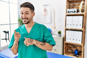 Canvas Print - Young physiotherapist man working at pain recovery clinic pointing to the back behind with hand and thumbs up, smiling confident