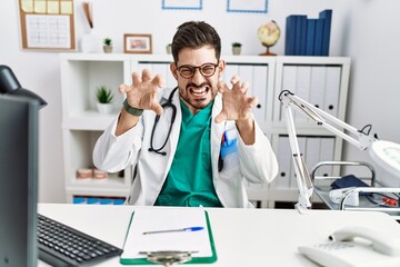 Poster - Young man with beard wearing doctor uniform and stethoscope at the clinic smiling funny doing claw gesture as cat, aggressive and sexy expression